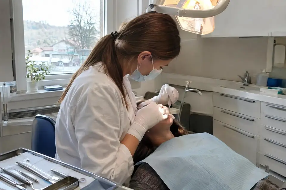Zobozdravnica z masko in rokavicami pregleduje pacientko, ki leži na zobozdravniškem stolu, ob njej pladenj z instrumenti.